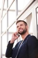 Good talk. Low angle view of confident young man in smart casual wear talking on the mobile phone and smiling while standing indoors photo