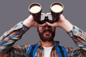 Exploring new places. Confident young bearded man carrying backpack and looking through binoculars while standing against grey background photo