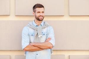 encantador guapo. apuesto joven con los brazos cruzados y sonriendo mientras está de pie frente a la pared texturizada al aire libre foto