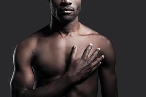It is in my heart. Cropped image of young shirtless African man holding his hand on heart while standing against grey background