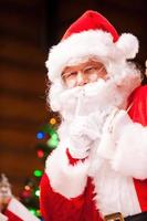 Shhh Traditional Santa Claus gesturing silence sign while carrying sack with presents and with Christmas Tree in the background photo