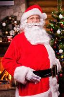 Ho Ho Ho Cheerful Santa Claus adjusting his belt and smiling with fireplace and Christmas Tree in the background photo