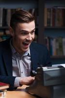 Just inspired. Happy young author typing something at the typewriter and keeping mouth open while sitting at his working place with bookshelf in the background photo