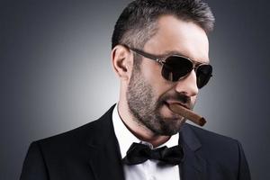 Bossy and self-confident. Portrait of handsome mature man in formalwear and sunglasses smoking cigar and looking away while standing against grey background photo