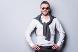 Confident in his perfect style. Handsome young man in smart casual wear and sunglasses looking at camera and smiling while holding hands on hip and standing against grey background photo