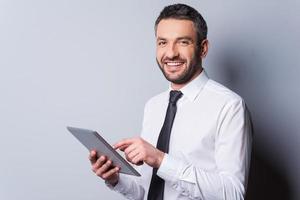 Feeling satisfied with his new gadget. Confident mature man in shirt and tie working on digital tablet and smiling while standing against grey background photo