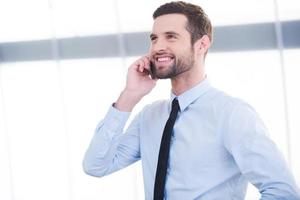 buena charla de negocios. joven empresario confiado hablando por teléfono móvil y sonriendo mientras está de pie en el interior foto