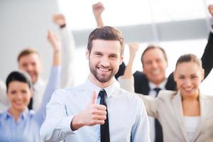 sintiéndose confiado en su equipo. hombre de negocios feliz mostrando su pulgar hacia arriba y sonriendo mientras sus colegas parados en el fondo foto