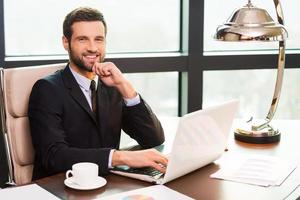 Confident business expert. Handsome young man in suit holding hand on chin and smiling while sitting at his working place photo