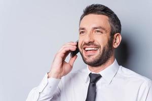 Great news Happy mature man in shirt and tie talking on the mobile phone and smiling while standing against grey background photo