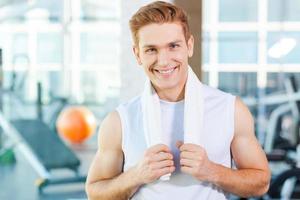 Strong and confident. Confident young muscular man carrying towel on shoulders and smiling while standing in gym photo