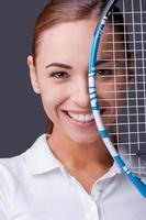 Pretty competitive.  Beautiful young women in sports clothes holding tennis racket in front of half of her face and looking at camera while standing against grey background photo