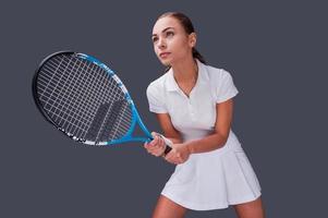 Ready to win. Low angle view of beautiful young women in sports clothes holding tennis racket and looking away while standing against grey background photo