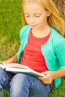 Reading her favorite book. Top view of cute little blond hair girl reading book while sitting on green grass and leaning at the tree photo