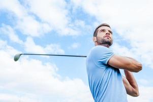 Professional golfer. Low angle view of young and confident golfer swinging his driver and looking away with blue sky as background photo