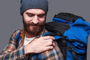 mochila demasiado pesada. frustrado joven barbudo ajustando su mochila mientras está de pie contra el fondo gris foto