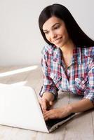 Spending time in the net. Beautiful young woman working on laptop and smiling while lying on the hardwood floor photo