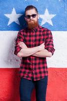 confiado y luciendo genial. apuesto joven barbudo con gafas de sol con los brazos cruzados y mirando a la cámara mientras se enfrenta a la bandera americana foto