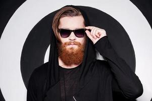 Mysterious bearded. Handsome young bearded man in sunglasses wearing hood and looking at camera while standing against stripped background photo