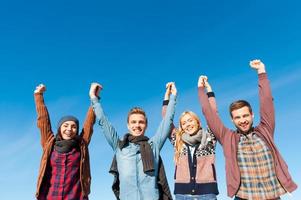 Forever young. Low angle view of four young happy people holding hands and raising them up with blue sky as background photo