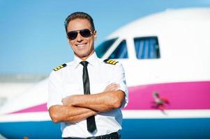 piloto confiado. piloto masculino confiado en uniforme con los brazos cruzados y sonriendo con un avión en el fondo foto