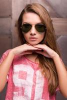 Casual beauty. Beautiful young woman in sunglasses holding hands on chin and looking at camera while standing against metal background photo