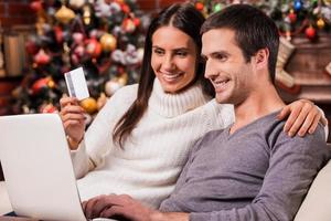 Taking advantages of Christmas sales. Beautiful young couple buying online while using computer together with Christmas Tree in the background photo