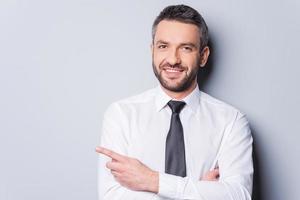 Pointing your product. Portrait of handsome mature man in shirt and tie pointing copy space and smiling while standing against grey background photo