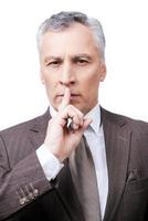 Keep my secret  Serious mature man in formalwear holding finger on lips and looking at camera while standing against white background photo