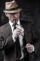 Money and power. Serious senior man in hat and suspenders smoking cigar and hiding money into the pocket while standing against dark background photo