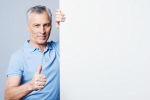 Confident at success. Handsome senior man showing thumb up and leaning at copy space while standing against grey background photo