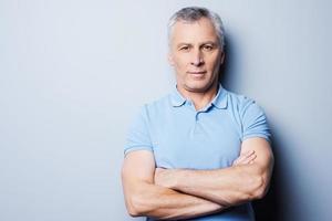 Confident mature man. Portrait of confident senior man in T-shirt looking at camera and smiling while keeping arms crossed and standing against grey background photo
