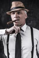 Bad news for you. Bossy senior man in hat and suspenders smoking cigar and holding finger on his neck while standing against dark background photo