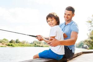 pescar juntos es divertido. alegre padre e hijo pescando juntos mientras el niño mira la cámara y sonríe foto