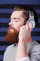 On the musical waves. Confident young bearded man in headphones standing against stripped background and listening to the music photo