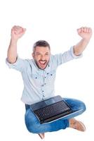 Top view of happy young man in casual wear keeping arms raised while sitting on the floor and with laptop while being isolated on white background photo