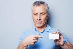 Call this number Handsome senior man pointing on a business card and smiling while standing against grey background photo