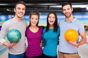 We love this game. Cheerful friends looking at camera and smiling while standing against bowling alleys photo