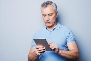 New gadget, the best assistant. Confident senior man working on his digital tablet while standing against grey background photo