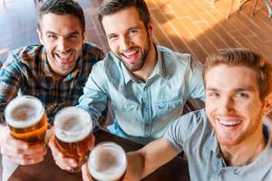 Top view of three happy young men in casual wear toasting with beer while sitting in bar together photo