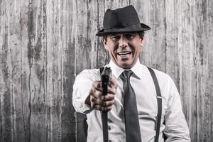 Say good bye Bossy senior man in gangster clothing stretching out hand with gun while standing against grey wall photo