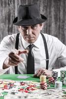 un anciano serio con camisa y tirantes sentado en la mesa de póquer y mirando la cámara con cartas con dinero y fichas de juego a su alrededor foto