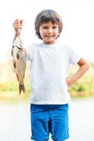 I catch it by myself Happy little boy carrying big fish on the hook and smiling while standing at the riverbank photo