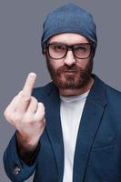 This is for you Handsome young bearded man showing hand with middle finger outstretched looking at camera while standing against grey background photo