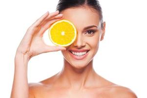 Great food for a healthy lifestyle. Beautiful young shirtless woman holding piece of orange in front of her eye while standing against white background photo