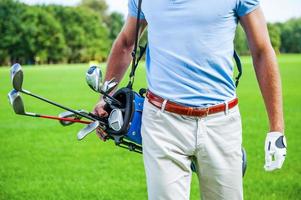 Ready to play. Close-up of male golfer carrying golf bag with drivers while walking by green grass photo