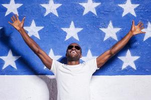 Welcome to USA Happy young African man in sunglasses raising his arms up and smiling while standing against American flag photo