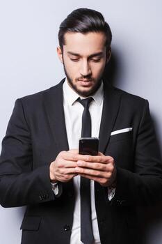 Working on the go. Confident young man in formalwear holding mobile phone and looking at it while standing against grey background photo