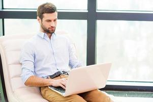 Working at home. Handsome young man working on laptop while sitting at the chair at home photo