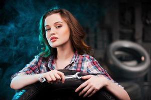Beautiful auto mechanic. Beautiful young woman holding work tool and looking at camera while leaning at the car tire in auto repair shop photo